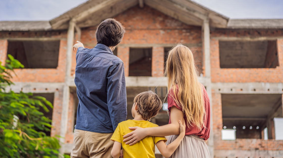Mutter, Vater und ihr kleiner Sohn stehen vor dem Rohbau ihres neuen Hauses und begutachten es. Der Vater zeigt nach links oben zum Haus, während die Mutter den Arm um die Schulter des Sohnes legt.