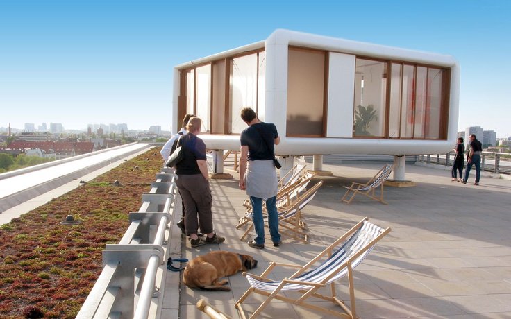 Drei Personen stehen vor einem modularen Haus, das auf einem Hausdach platziert ist