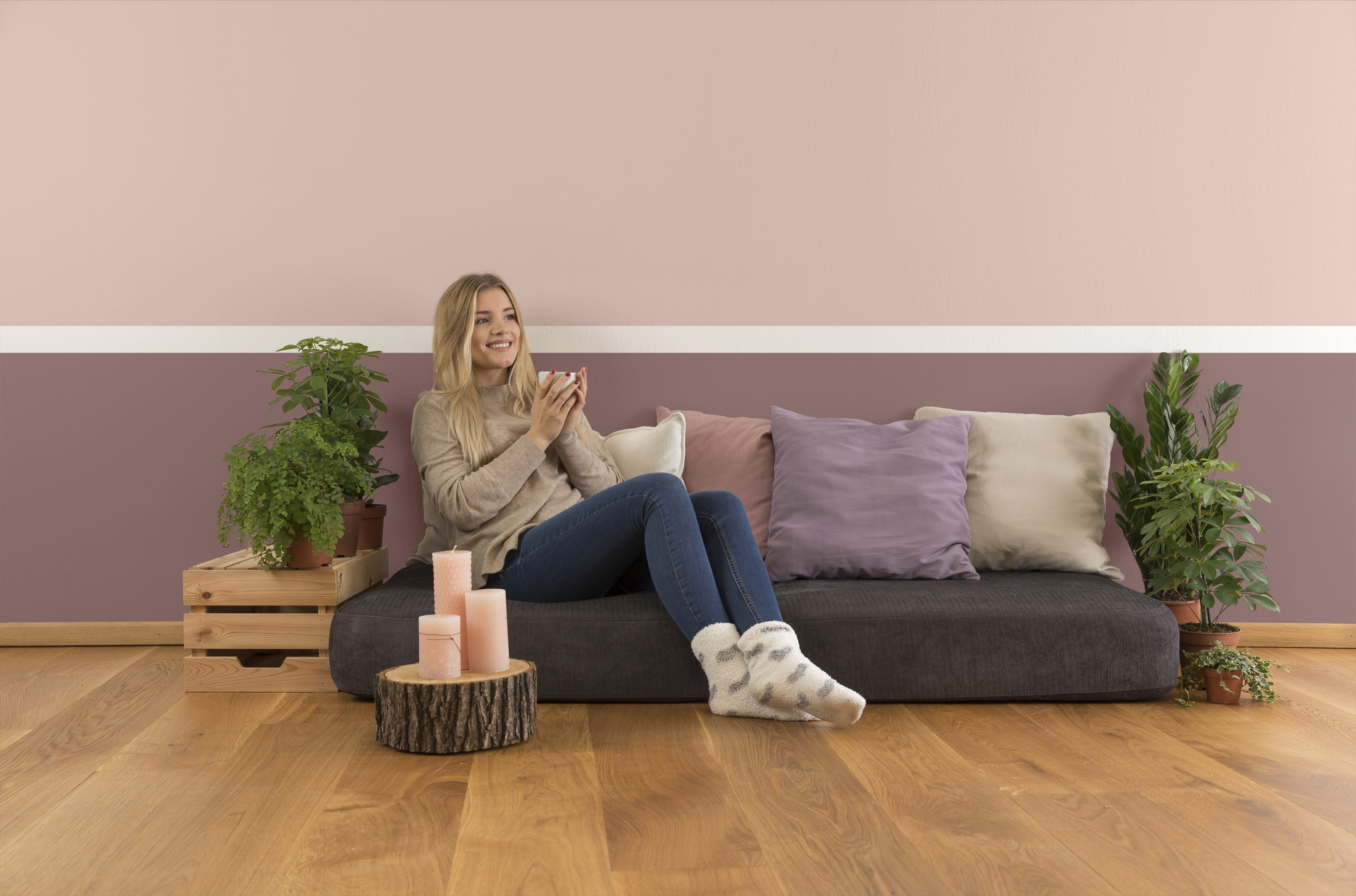 Junge Frau sitzt mit Teetasse auf brauner Matratze vor rosa und Brombeerfarben gestrichener Wand