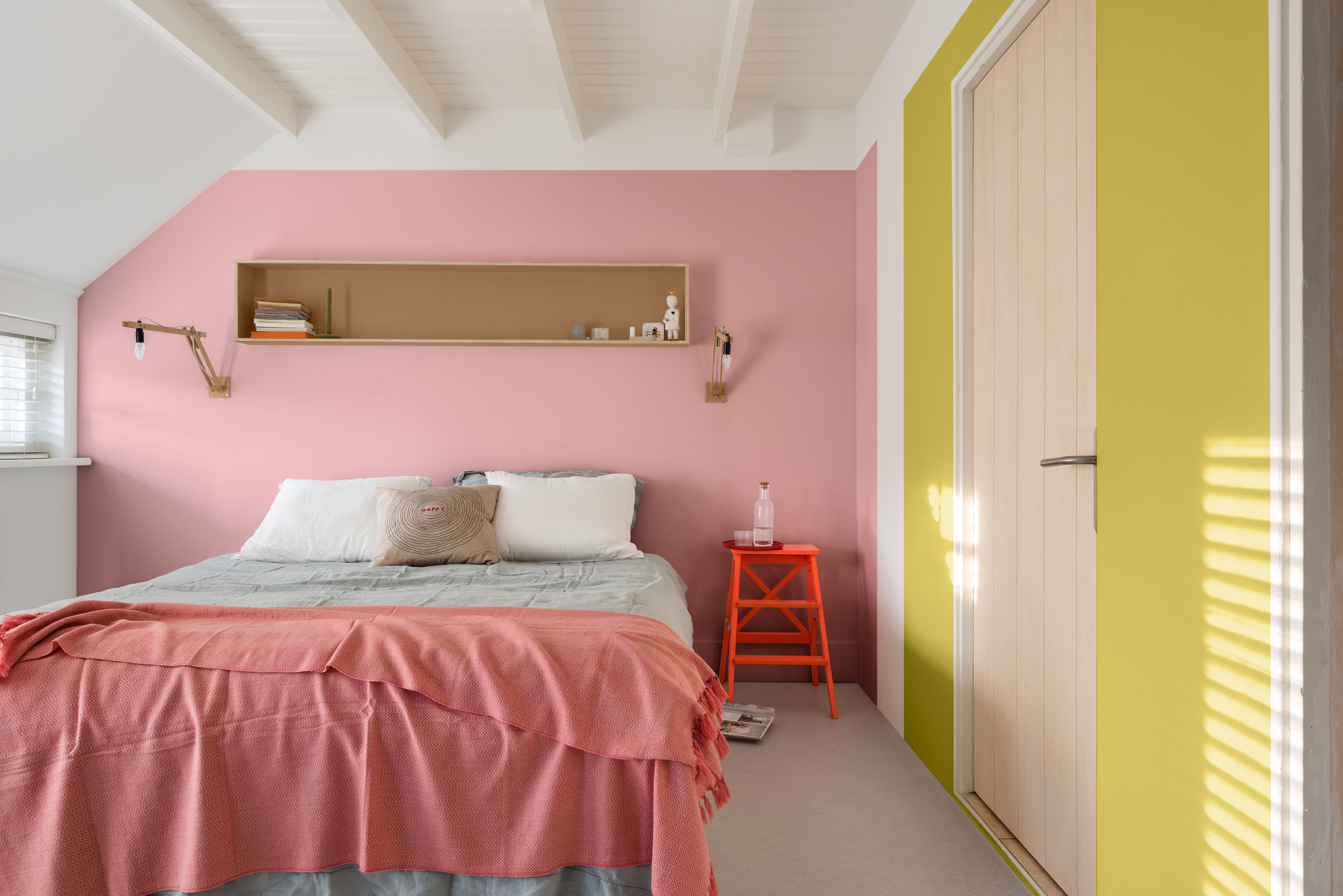 Schlafzimmer mit Bett vor rosa und gelb gestrichener Wand und rotem Hocker als Nachttisch.