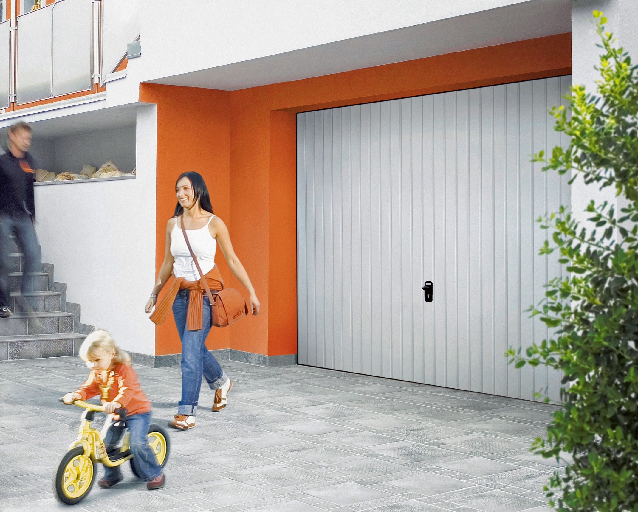 Kind mit Laufrad und Mutter vor einer Garage.