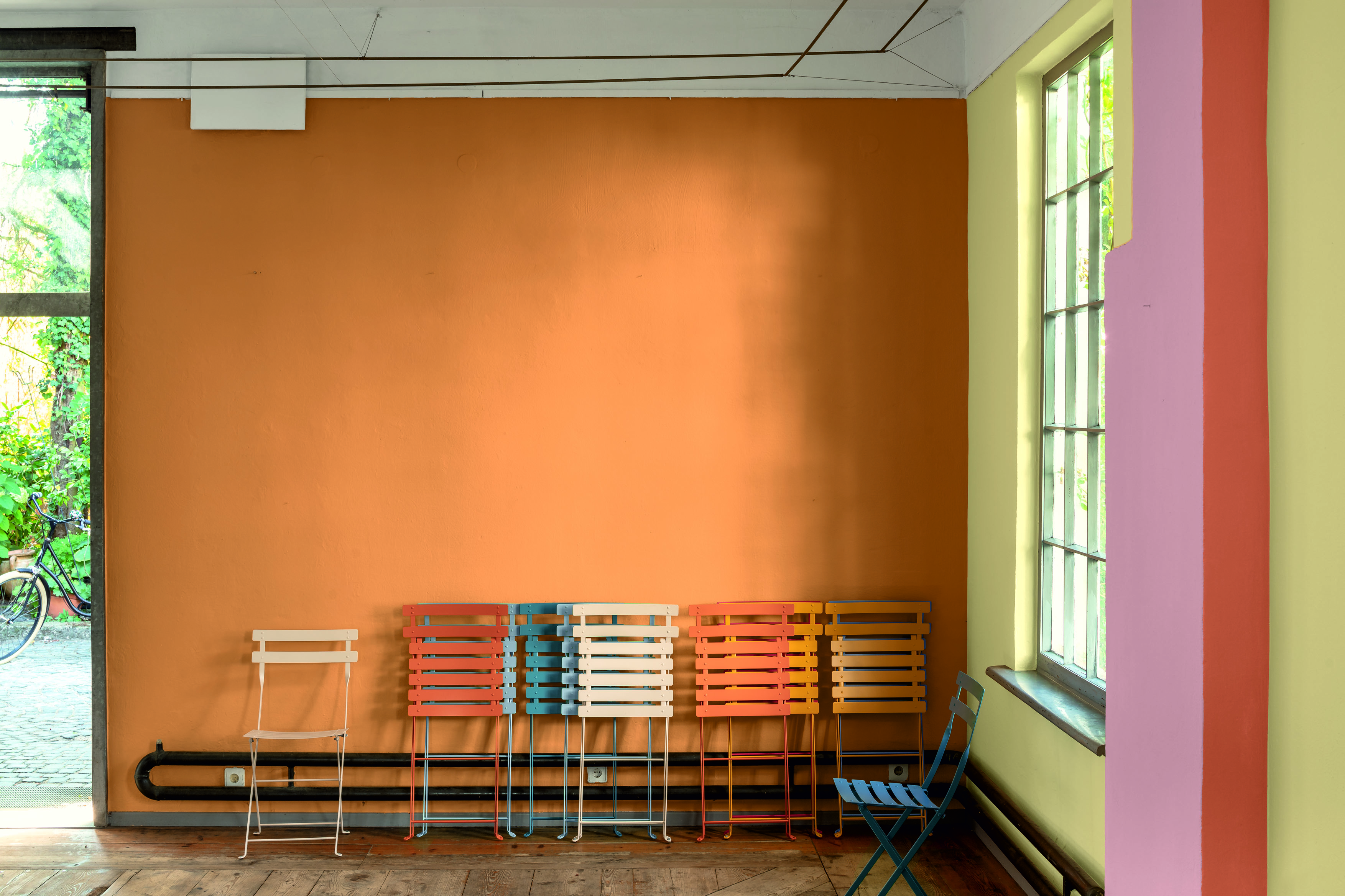 Klappbare Gartenstühle in verschiedenen Farben lehnen neben einem hohen Fesnter an einer Orange gestrichenen Wand.