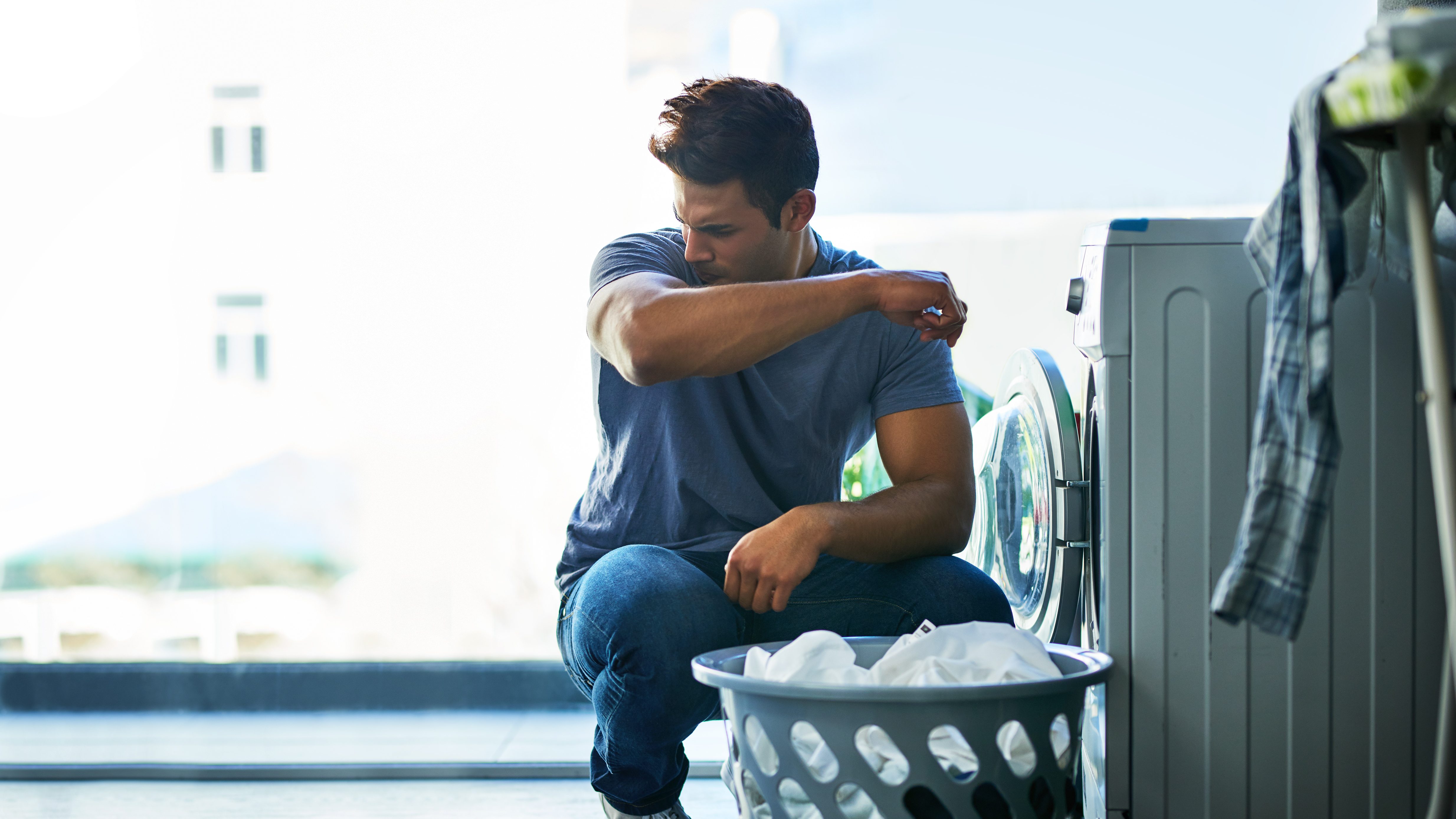 Mann sitzt vor Waschmaschine und riecht an T-Shirt