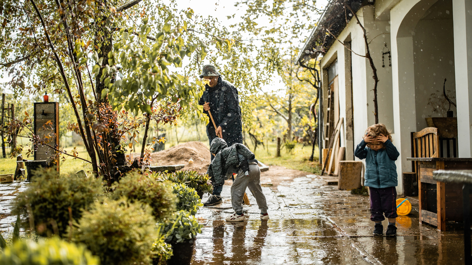 Mann steht mit zwei Kindern vor dem Haus im Regen.
