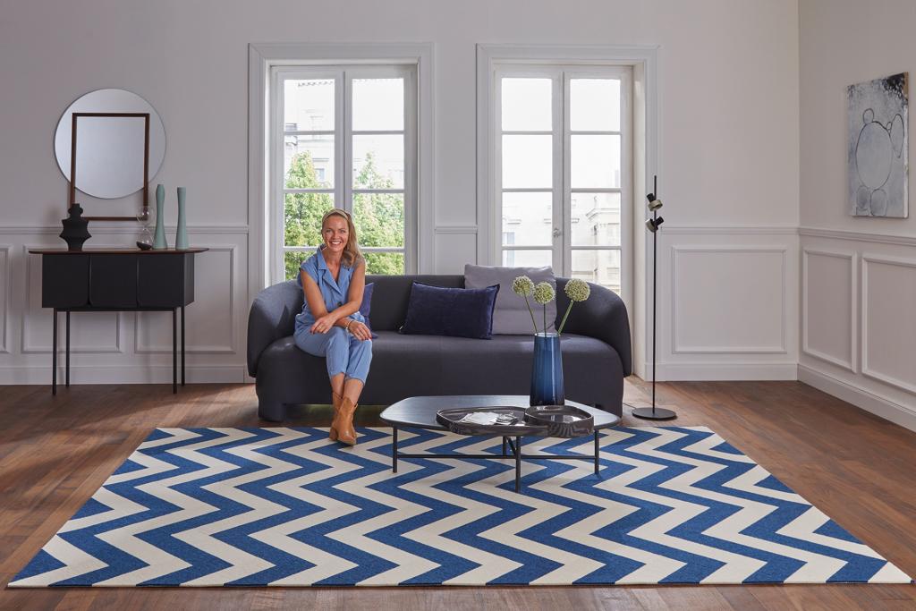 TV-Moderatorin Eva Brenner sitzt auf einem Sofa in einem Wohnzimmer, in dem ein Tretford Designteppich INTERART, Design Amsterdam, ausgelegt ist.