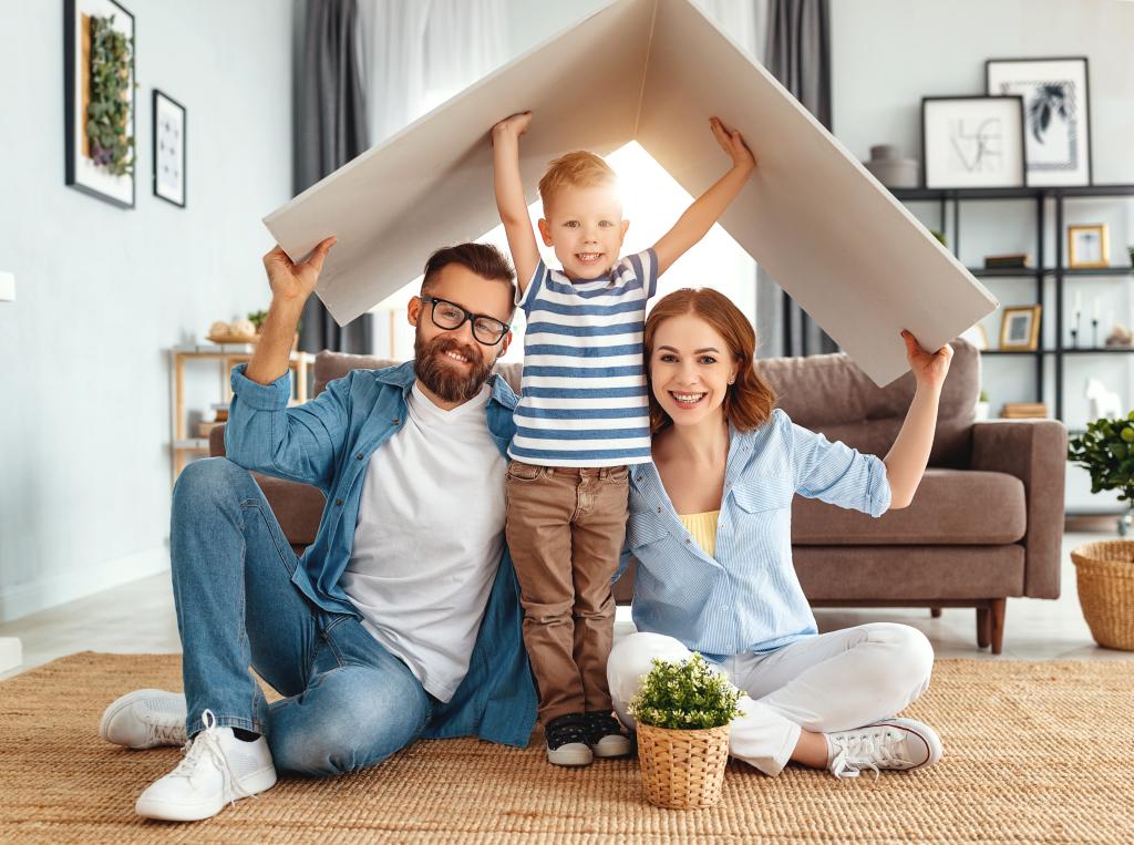 Eine junge Familie sitzt lächelnd auf dem Boden ihres Wohnzimmers, während das Kind steht. Gemeinsam halten sie sich ein Stück Pappe in Form eines Hausdachs über die Köpfe.