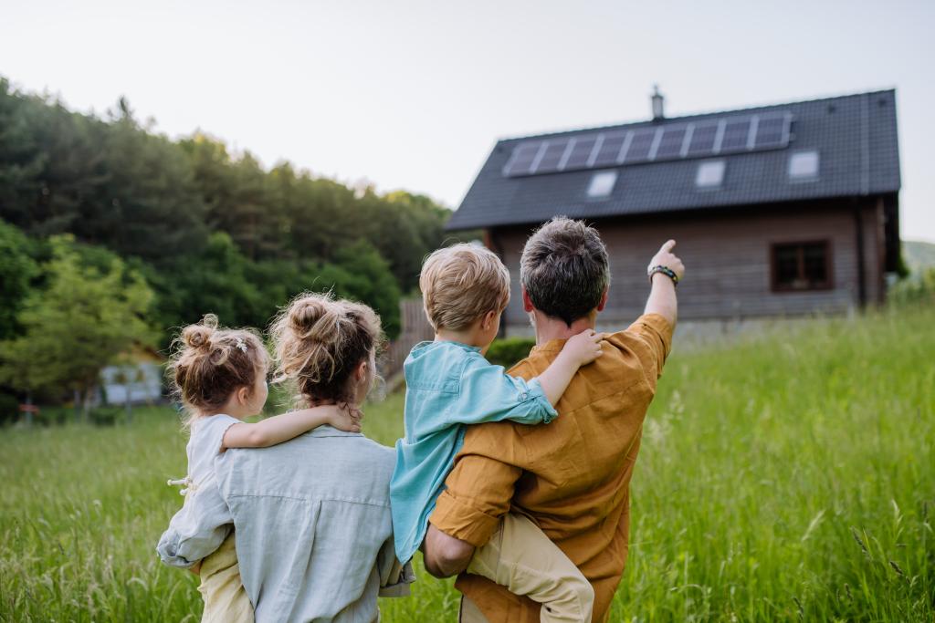 Junge Eltern haben jeweils eines ihrer kleinen Kinder auf dem Arm und begutachten ein freistehendes Einfamilienhaus, auf das der Vater zeigt.