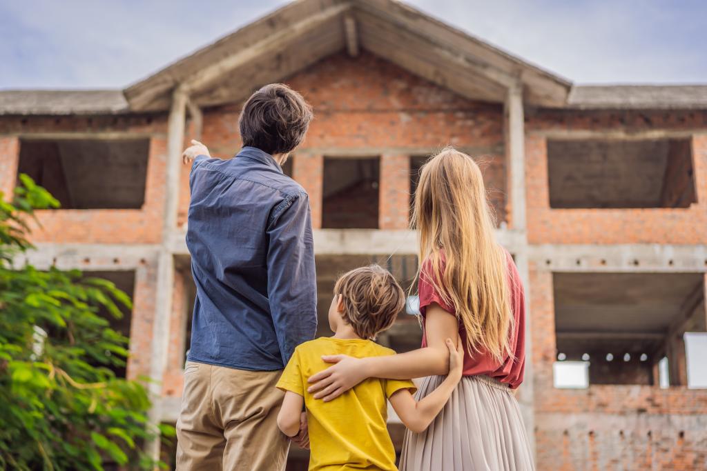 Mutter, Vater und ihr kleiner Sohn stehen vor dem Rohbau ihres neuen Hauses und begutachten es. Der Vater zeigt nach links oben zum Haus, während die Mutter den Arm um die Schulter des Sohnes legt.