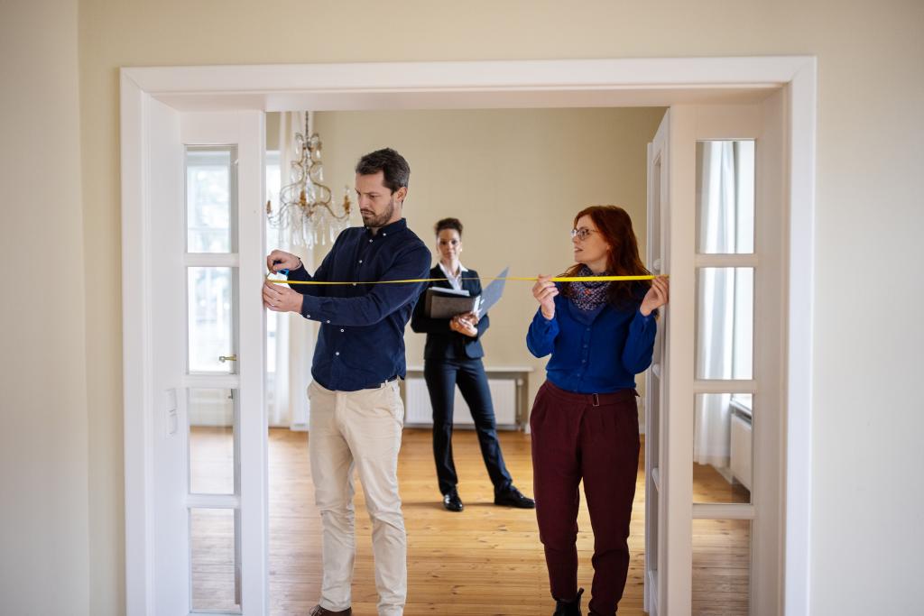 Ein Mann und eine Frau vermessen mit einem Maßband die Breite einer Tür. Im Hintergrund steht die Immobilienmaklerin mit einer Unterlagenmappe und steht für weitere Fragen zur Verfügung.