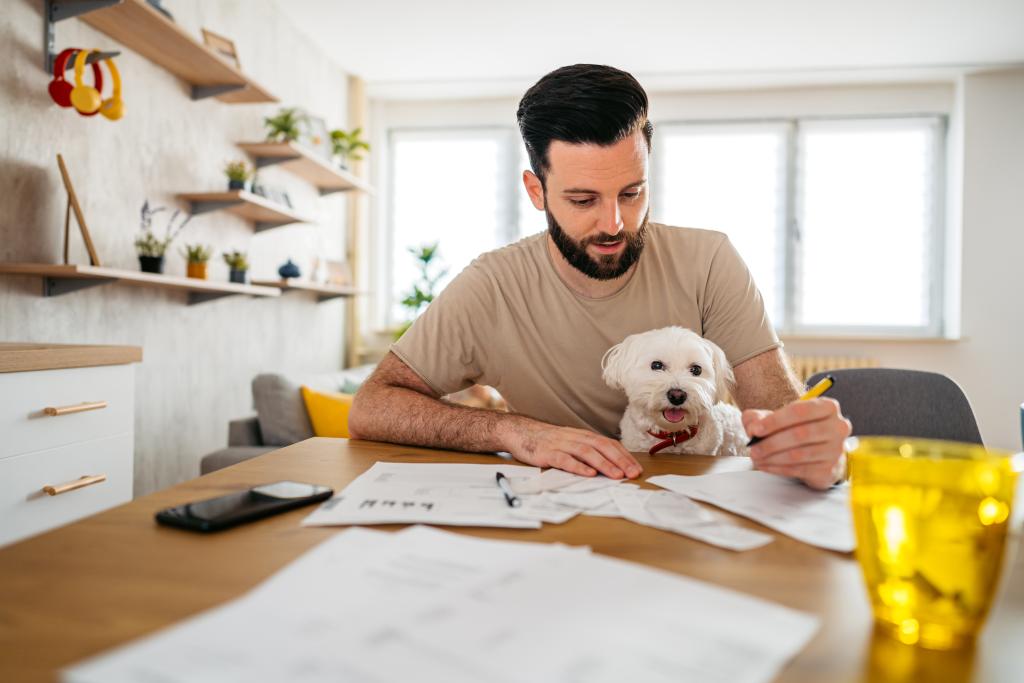 Ein Mann sitzt mit Hund auf dem Schoß am Tisch und überprüft in Unterlagen Finanzen.