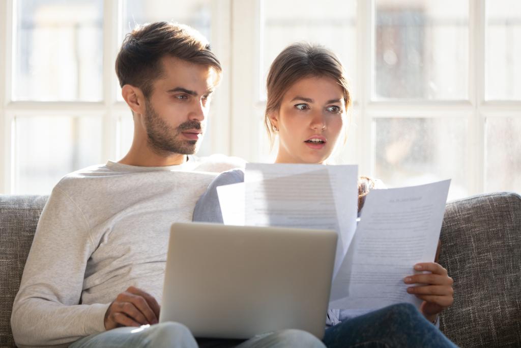 Junges Paar sitzt mit einem Laptop auf dem Sofa und blickt erschrocken auf Unterlagen