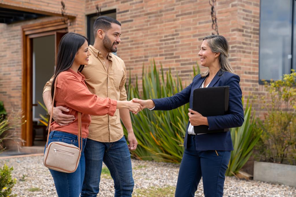 Paar und Maklerin schütteln sich die Hände vor einem Haus.