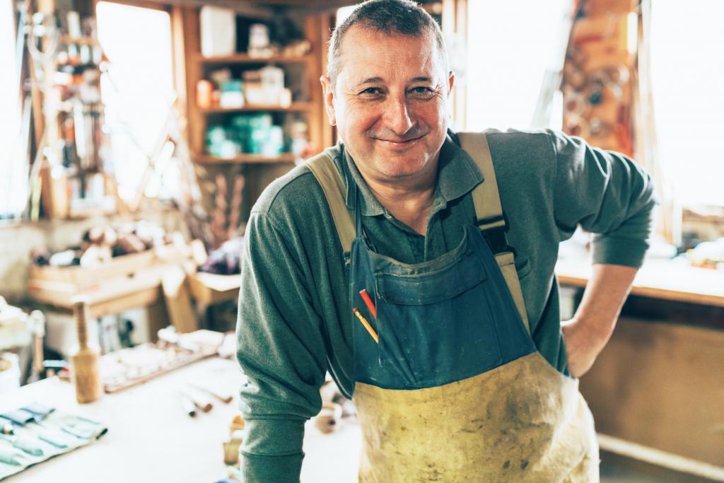 Ein Handwerker mit Arbeitsschürze steht in seiner Werkstatt und lächelt.