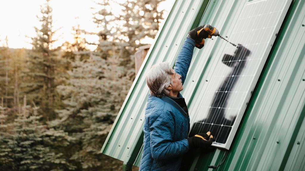 Grauhaariger Mann schraubt Solarpanel am Dach an.