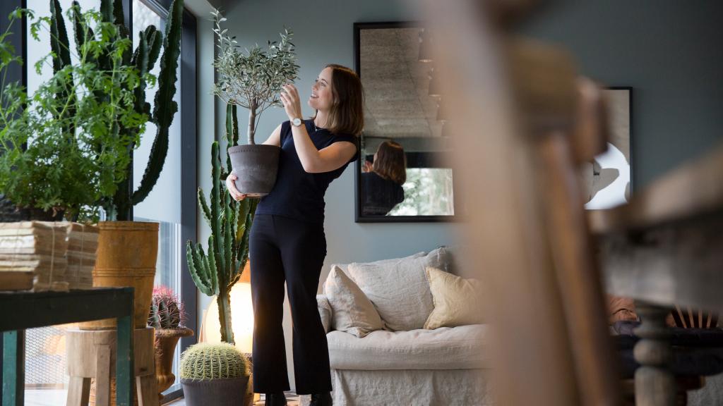 Frau hält kleines Bäumchen im Kübel in einem mit Pflanzen gefüllten Zimmer