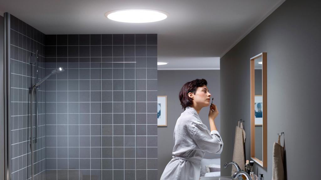 Eine Frau im Bademantel schminkt sich im Bad, während durch einen Tageslichtspot Licht fällt.